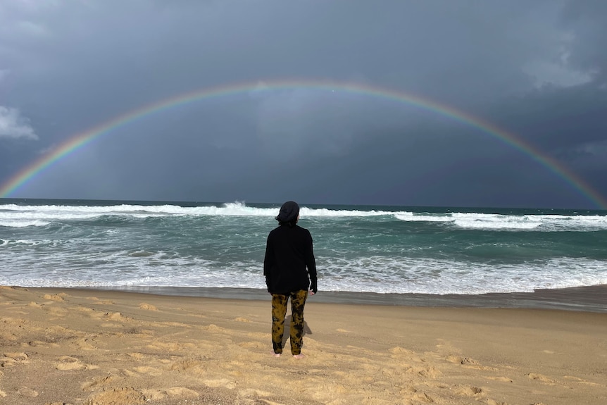 A man stands on a beach looking out to sea with a rainbow from edge to edge of the photo.