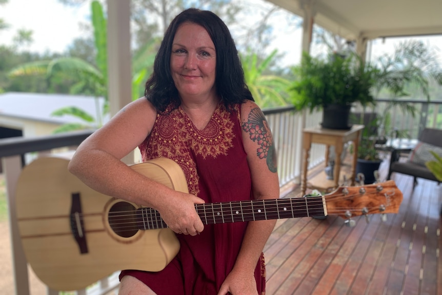 A woman wearing a red patterned top sits on a chair on a deck, holding a light-coloured guitar and smiling.