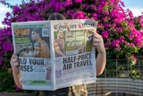 A woman holds up a newspaper in front of her face in front of a bush of pink flowers.