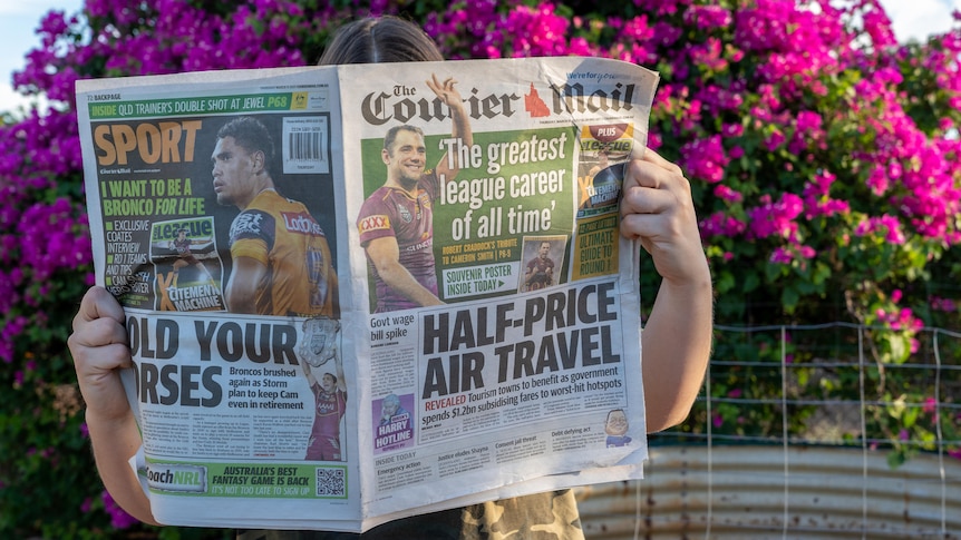 A woman holds up a newspaper in front of her face in front of a bush of pink flowers.