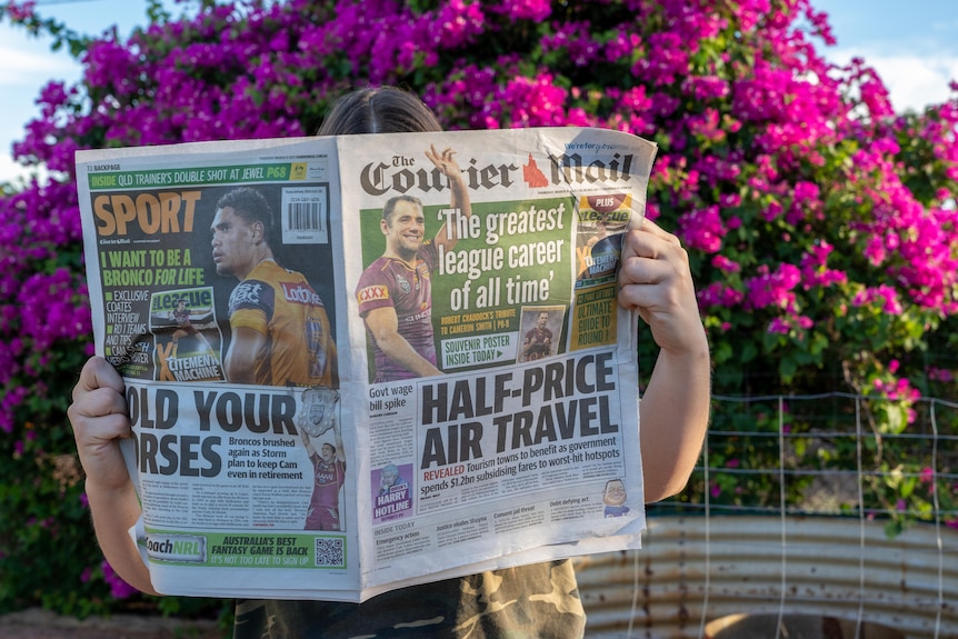 A woman holds up a newspaper in front of her face in front of her face in front of a bush of pink flowers.