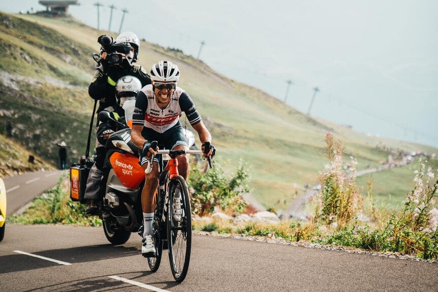 Richie Porte riding his bike up a hill with a photographer behind on a motorcycle.