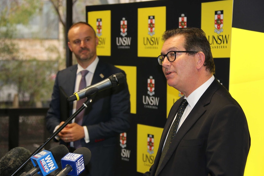 A middle-aged man with black rimmed glasses speaks into a microphone at a press conference