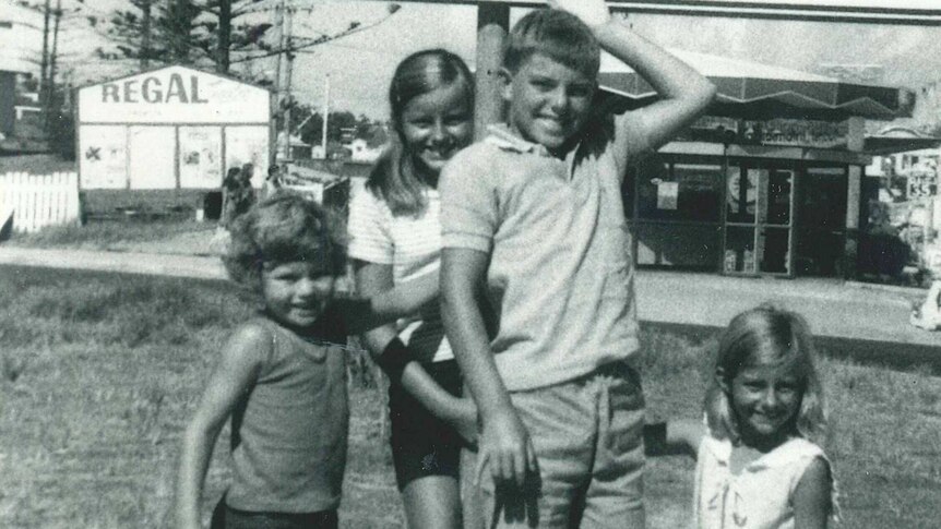 A black and white photo of four kids at the state border, pointing to the sign above them