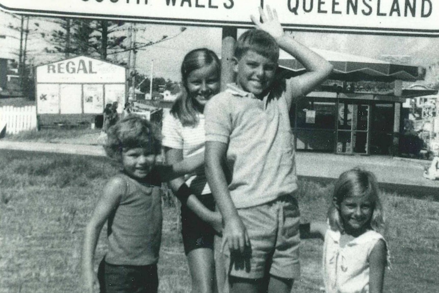 A black and white photo of four kids at the state border, pointing to the sign above them
