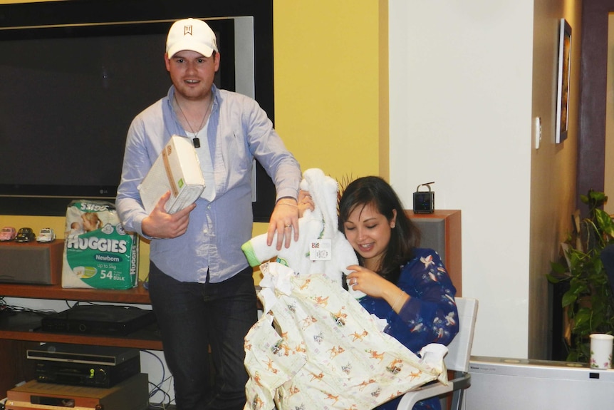 A man standing  has his hand on a woman's shoulder, who is sitting, opening presents for a baby to depict story of stillbirth.