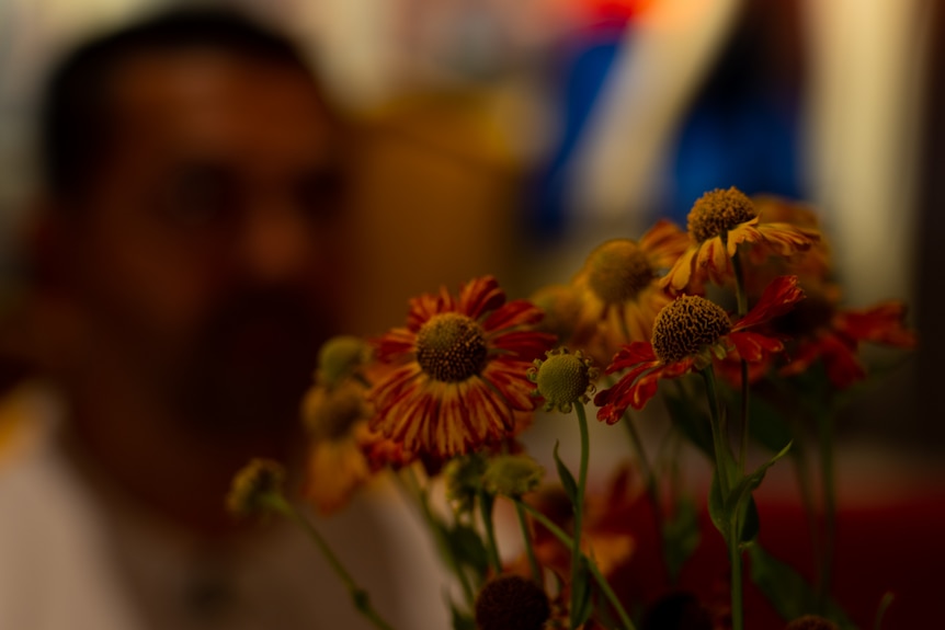 Flowers sit in the foreground in front of a man with a face out of focus.
