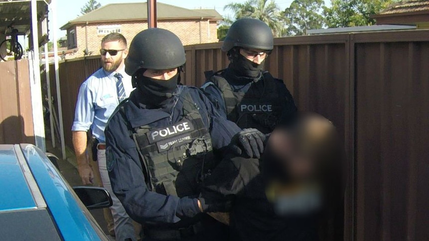 Two police in riot gear lead a man, blurred out, down a path. Behind them is a plainclothes detective.