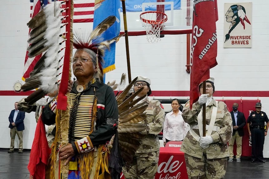 Native Americans dressed in traditional clothing lead a ceremony in a school gymnasium.