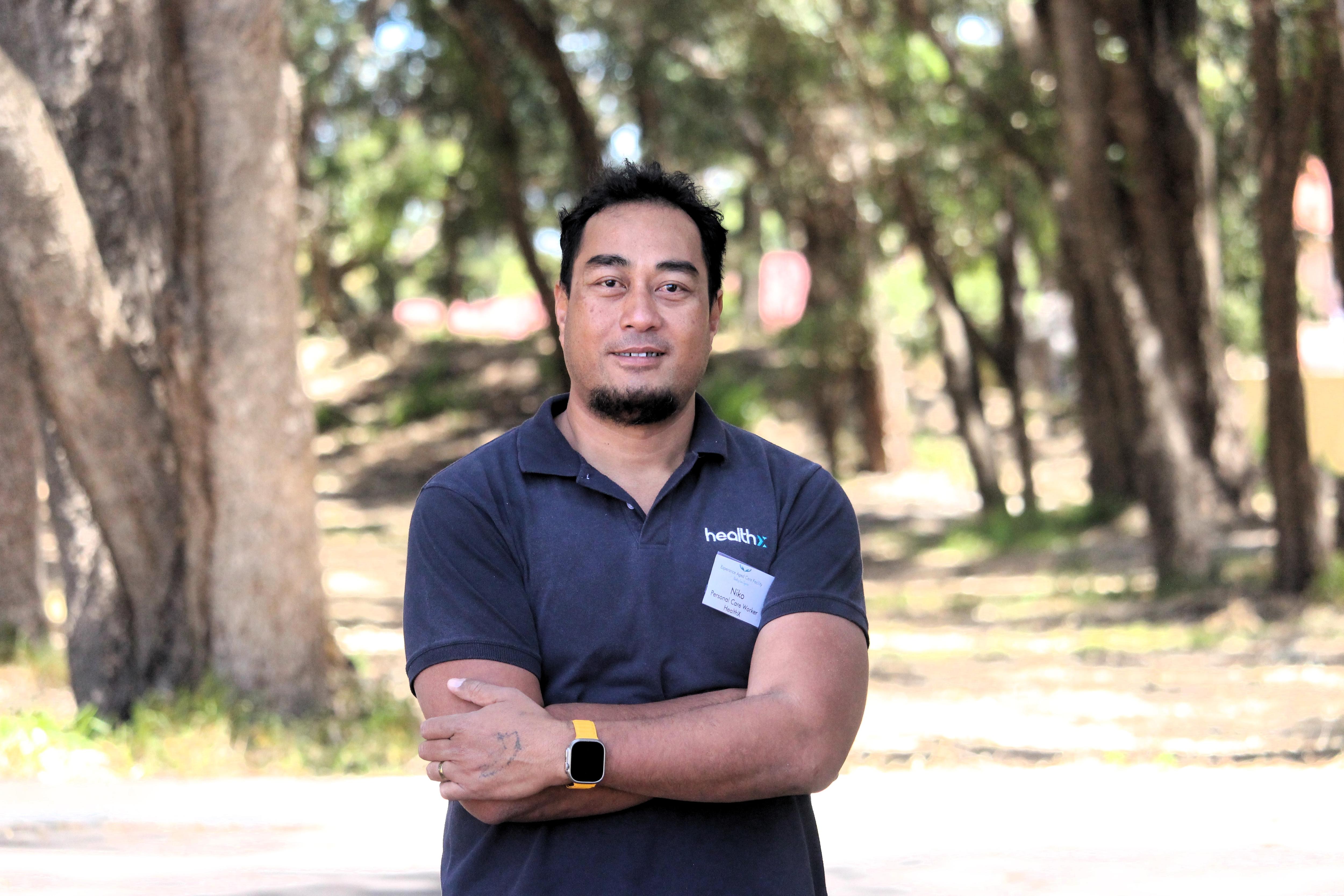 He stands near some trees with his arms folded