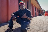 an aboriginal man wearing a beanie squatting in the sun in front of a red building