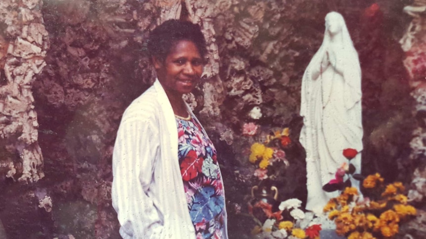 A smiling woman stands against a rocky outcrop next to an alcove with a catholic statue of Mary surrounded by flowers.