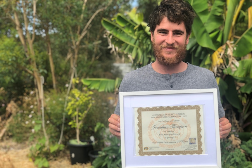 Young man with a beard holding up a framed certificate in front of a banana tree and garden.