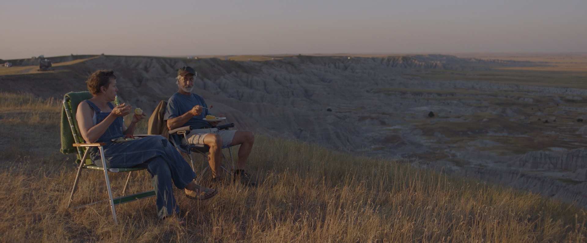 Late afternoon shot of older woman and man in camper chairs overlooking flat grassland, eating from plates in their laps.