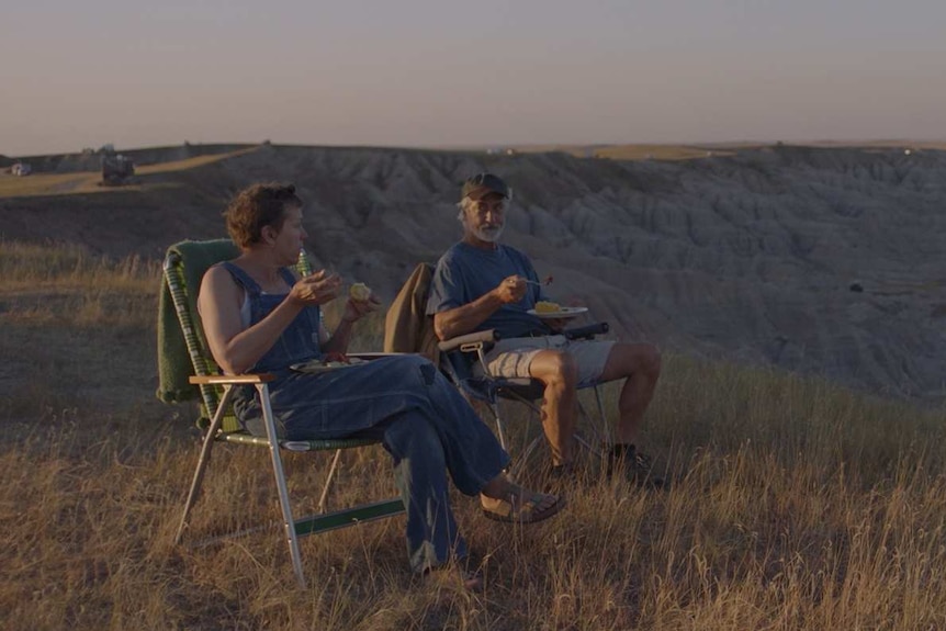 Late afternoon shot of older woman and man in camper chairs overlooking flat grassland, eating from plates in their laps.