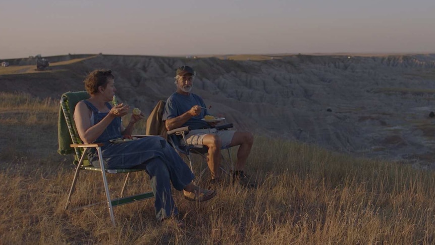 Late afternoon shot of older woman and man in camper chairs overlooking flat grassland, eating from plates in their laps.