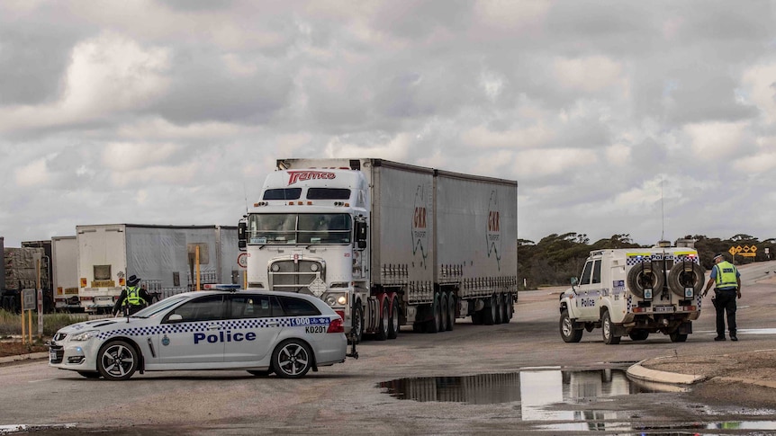 Police vehicles parked up as part of a road block at the WA border as police officers walk towards a parked truck.