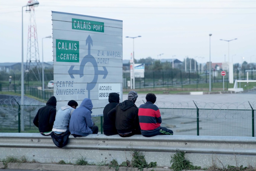 Kids sit in front of a big road sign with directions to Calais