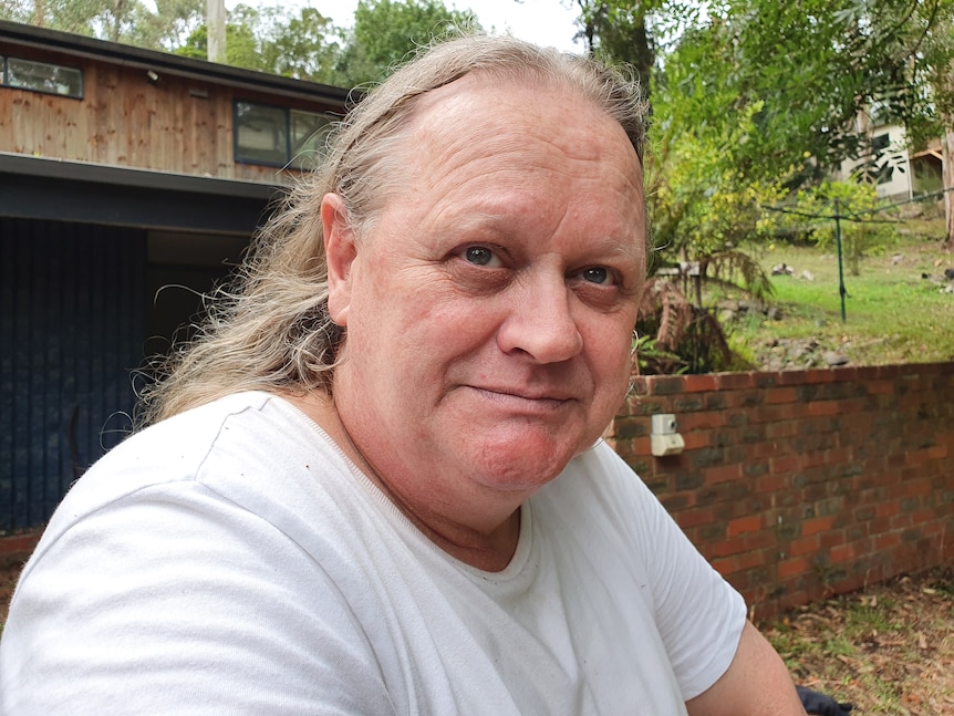 A middle-aged man with long hair sits in a backyard with a serious facial expression.