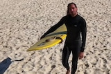 A long-haired man in a black wetsuit stands on the sand, holding a long yellow surfboard with a snake on the front of it.