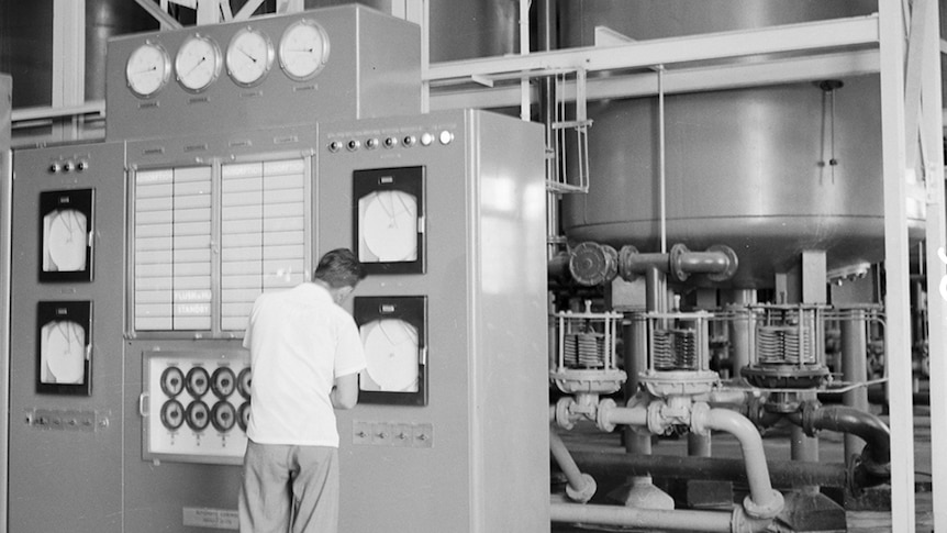A black and whitye photo of a man checking readings on a machine with pipes in the background.