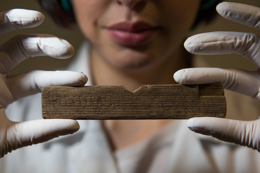 A woman holds up a small fragment of wood with faint words carved into it