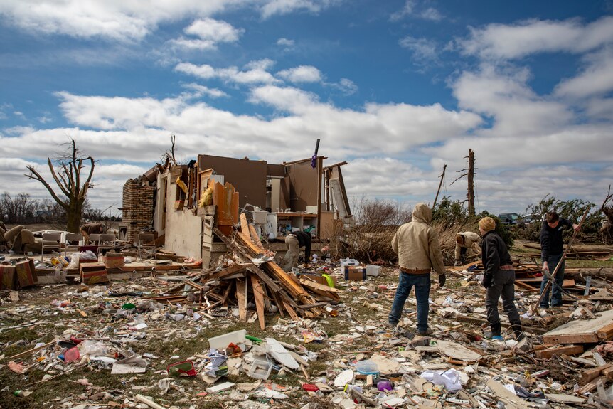 People look at a house that has lost its roof as household objects and debris surround them.