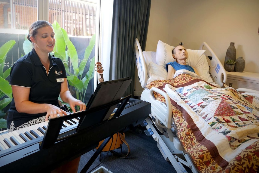 A woman plays piano keyboard next to a sick man in a hospital bed.