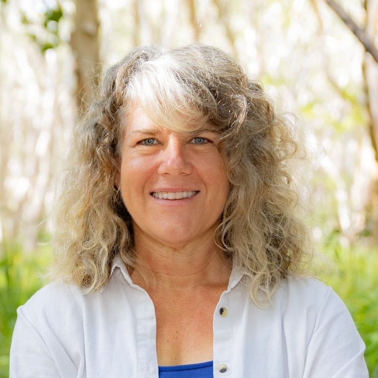 A smiling woman wearing a white shirt stands in a grassy, wooded area.