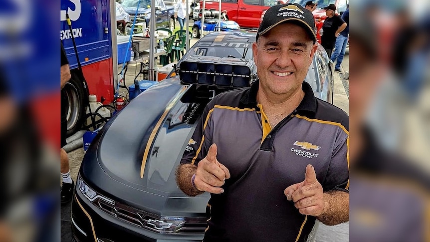 A 50's man in racing garb smiles and points two fingers in front of a car in garage