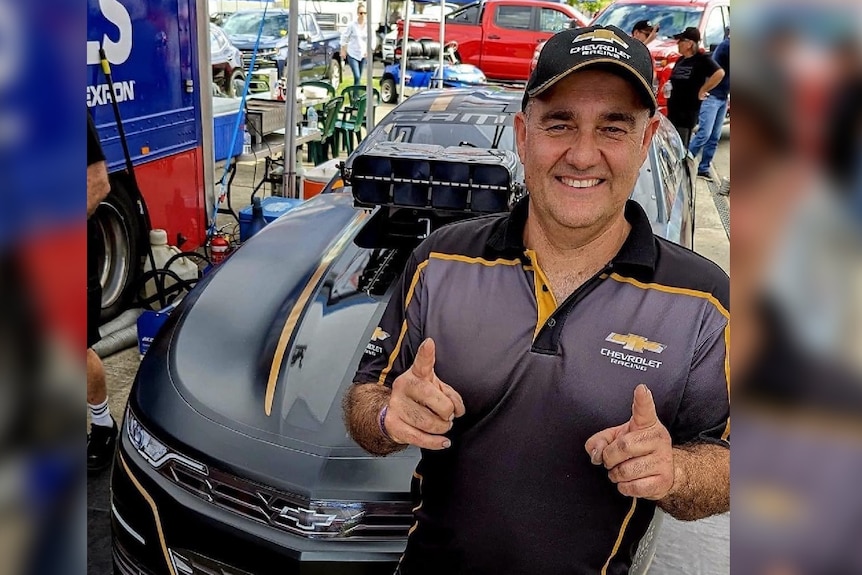 A man in his 50s smiles and points in front of a drag racing car.