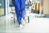 A doctor in blue scrubs walks along a hospital hall pushing a hospital bed in front of them