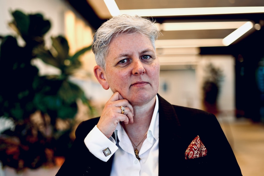 A womna with short grey hair, wearing a dark suit jacket over a whit sift, looks intently into the camera.