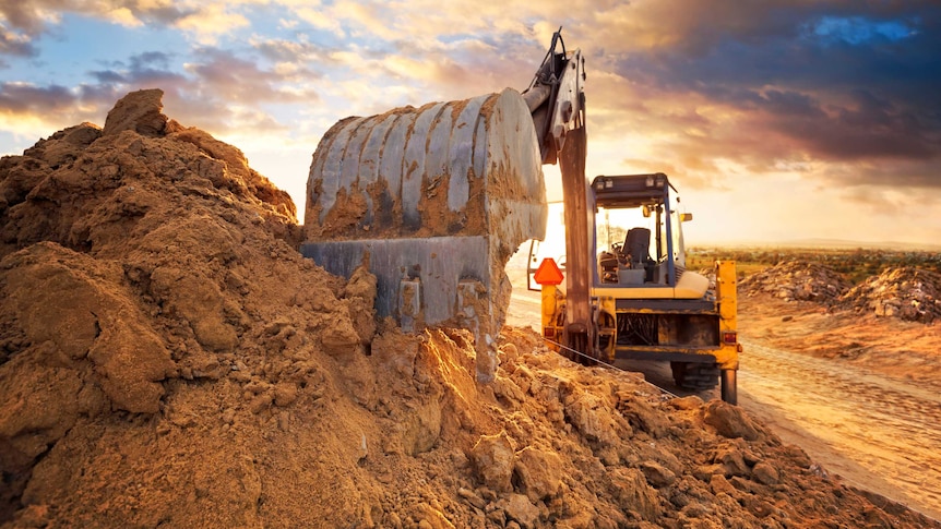 Under a colourful, cloudy sky, a large yellow digger digs at a huge pile of sand. Flattened dirt road is in the background.