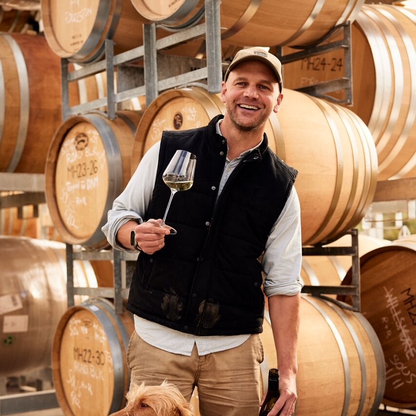 A man standing with a dog in front of wine barrels. The man is holding a glass and bottle of white wine.