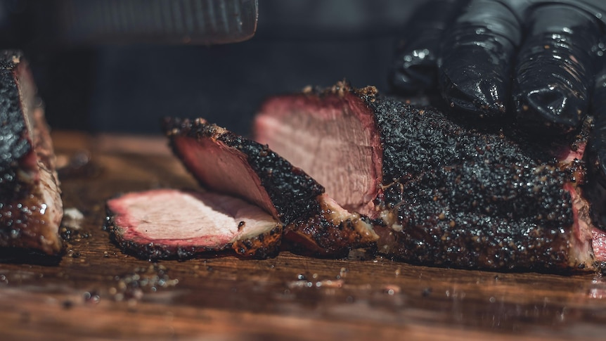 A close up of a thick piece of meat with a crusty charred black exterior, pink brown inside, cut into slices. 