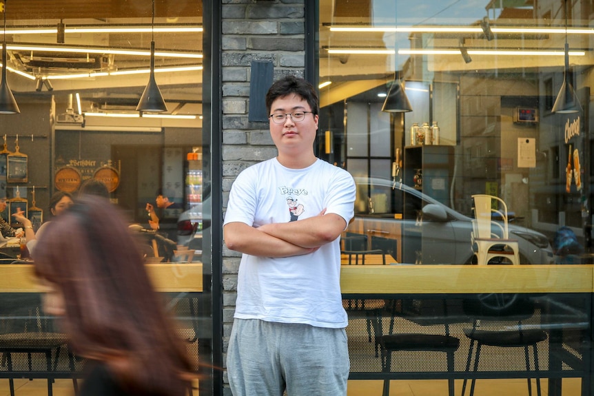 A South Korean man in a Popeye t-shirt stands in front of a cafe.