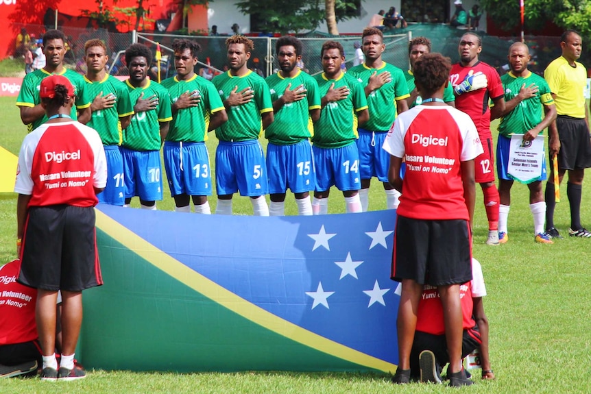 The Solomon Islands national team sings the national anthem before a game with the Solomons flag in the foreground.