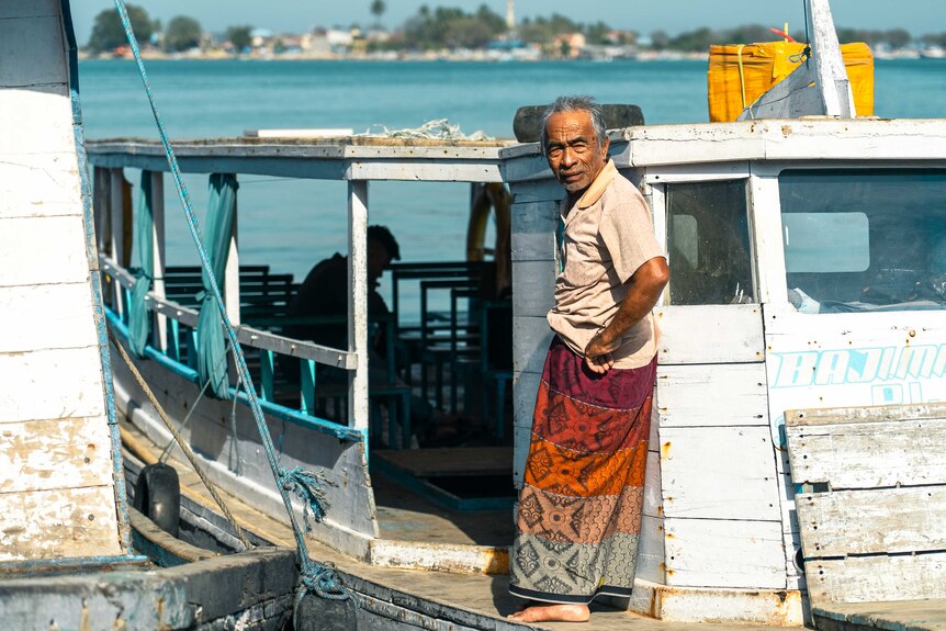 A fisherman in a sarong watches on, standing on a boat in the port of Makassar.