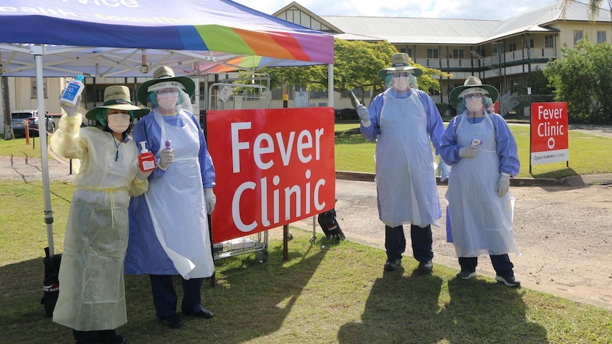 Four people in nursing gowns, smocks and face masks holding hand sanitiser in front of a tent.