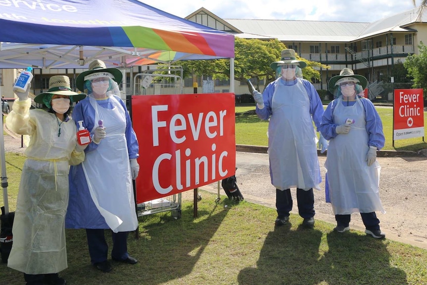 Four people in nursing gowns, smocks and face masks holding hand sanitiser in front of a tent.