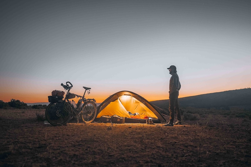 A man stands next to a tent and bicycle at sunset.