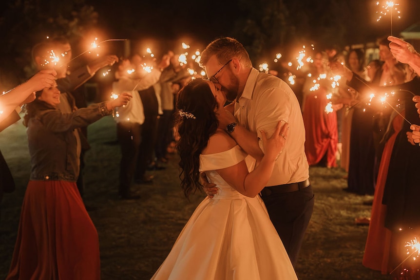 A bride kisses her groom among a crowd of people waving sparklers.