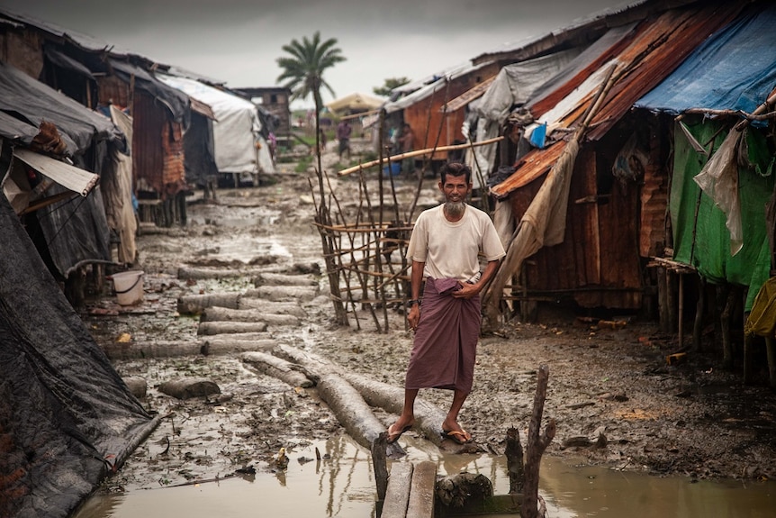 A man wearing a longyi balances on logs to walk amid mud and puddles between rows of tents in a refugee camp.