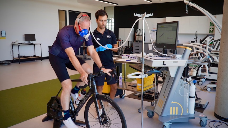 A scientist stands next to a cyclist on a stationary bike, looking at data, while the rider wears an oxygen mask