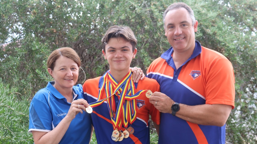 A woman, teenager and man standing in front of a bush holding a bunch of medals around the teenager's neck.