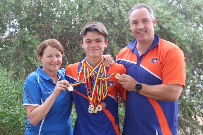 A woman, teenager and man standing in front of a bush holding a bunch of medals around the teenager's neck.