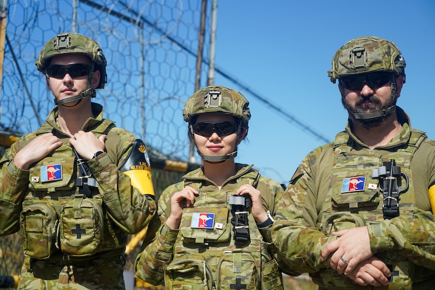 Two men and a woman wearing sunglasses and army fatigues stand straight, staring ahead.