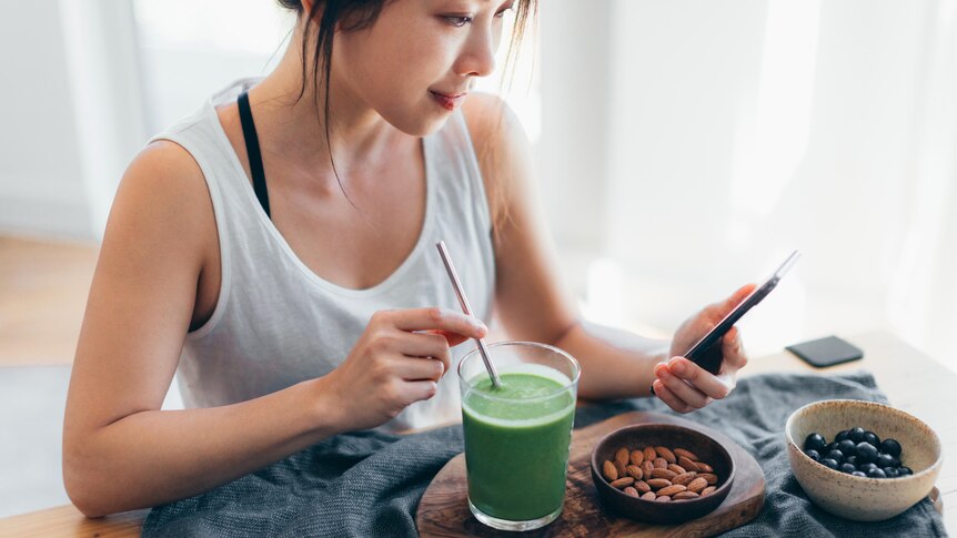 A woman looks at her phone while stirring a green smoothie. A bowl of almonds and a bowl of blueberries are also in front of h