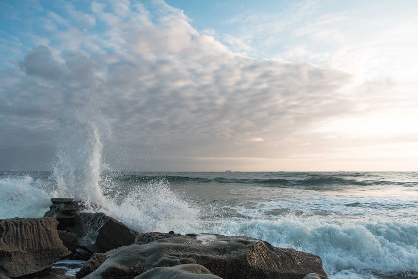 Fluffy white clouds with waves crashing on a rock formation.